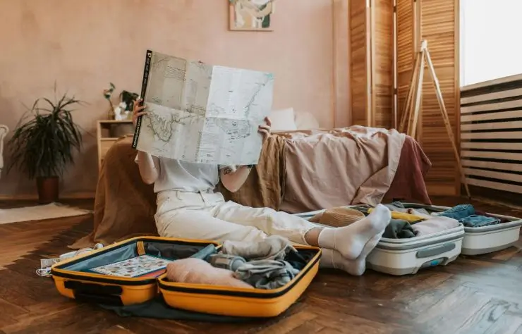 mujer sentada en el suelo recostada a la cama mirando un mapa de papel con maletas a los lados.